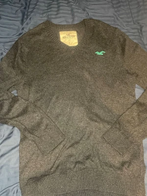 Mörkgrå stickad tröja Hollister - Mörkgrå stickad tröja från Hollister med V-ringning och ribbade muddar vid ärmslut och nederkant. Tröjan har en liten turkos Hollister-logga broderad på bröstet. Perfekt för lager på lager och enkel att matcha med jeans.