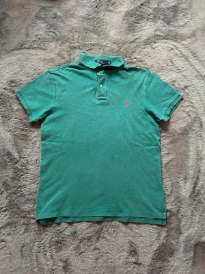 Grön pikétröja från Ralph Lauren - Klassisk grön pikétröja från Ralph Lauren med korta ärmar och krage. Tröjan har två knappar framtill och den ikoniska rosa logotypen broderad på bröstet. Tillverkad i mjuk bomull som känns skön mot huden. Perfekt för en avslappnad men ändå stilren look.