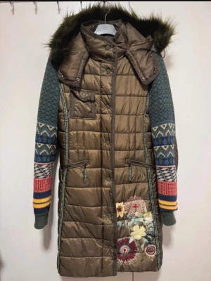 Desigual quiltad dunjacka med mönstrade ärmar - Unik lång dunjacka från Desigual i olivgrönt med quiltad kropp och stickade, mönstrade ärmar i blått, grönt, rött och gult. Jackan har broderade blommor, avtagbar huva med fuskpäls och färgglatt blommigt innerfoder. Två dragkedjefickor och snygga detaljer längs sidorna.