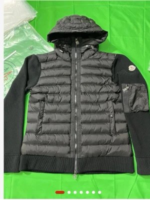 Svart dunjacka med huva från Moncler - Snygg svart dunjacka från Moncler med huva och ribbade stickade ärmar. Jackan har dragkedja framtill, två sidofickor med dragkedja och en extra ficka på ärmen. Klassisk Moncler-logga på vänster ärm. Perfekt för kyliga dagar.