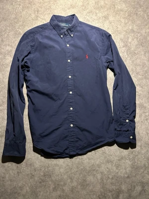 Marinblå skjorta från Polo Ralph Lauren - Klassisk marinblå skjorta från Polo Ralph Lauren i slim fit. Skjortan har knappar hela vägen, button-down krage och den ikoniska röda loggan broderad på bröstet. Tillverkad i mjuk bomull och långärmad modell. Passar som en S