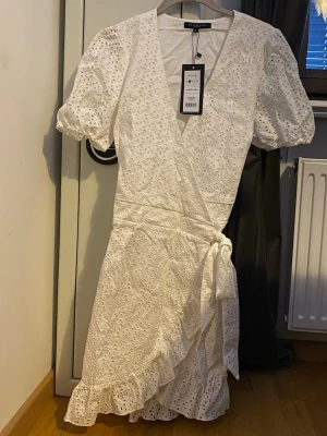 Vit omlottklänning från My Jewellery - Kan sänka i pris vid snabb affär! Superfin vit omlottklänning från My Jewellery med broderade detaljer och volangkant. Klänningen har korta puffärmar, knyt i midjan och v-ringning. Perfekt för sommarens alla tillfällen och eller student.