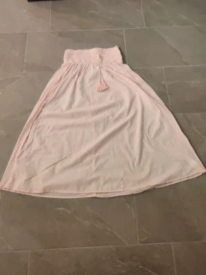 Ljusrosa långkjol med knytband - Söt ljusrosa långkjol med smockad midja och dekorativt knytband med tofsar. Kjolen har en flowy passform och är perfekt för varma dagar. Materialet känns mjukt och lätt mot huden. Den är i mycket bra skick men kan vara lite skrynklig då jag är usel på att stryka. Går även att vika ner vid midjan och är i storlek 158/164. 