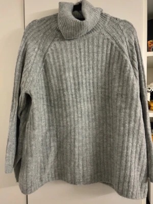 Grå ribbstickad polotröja - Mysig grå ribbstickad polotröja med lång ärm och hög krage. Tröjan har en avslappnad passform och är perfekt för kalla dagar. Materialet ser ut att vara mjukt och värmande, troligen ull- eller ullblandning. Enkel och stilren design som passar till det mesta.