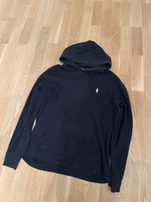 Polo Ralph Lauren - Säljer en marinblå från Polo Ralph Lauren med klassisk huva och stor ficka framtill. Tröjan har det ikoniska vita Polo-märket broderat på bröstet och är gjord i ett mjukt, strukturerat bomullstyg. Perfekt för en avslappnad och stilren look.