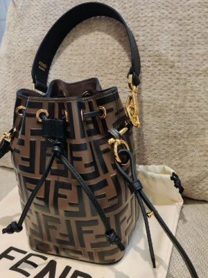 Fendi Monogram handväska i skinn - Säljer en brun och svart handväska från Fendi med ikoniskt FF-monogram. Väskan är inte i skinn och har guldfärgade metalldetaljer samt snörning upptill. Praktiskt axelband och exklusiv design som sticker ut.