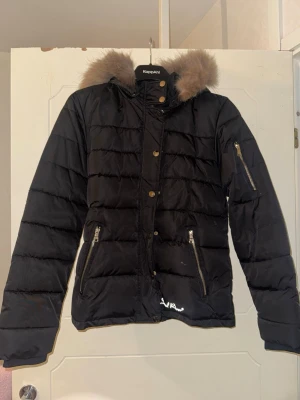 Svart dunjacka Soul River med fuskpäls - Svart quiltad dunjacka från Soul River med avtagbar huva och fuskpäls. Jackan har dragkedjor i guld, fickor fram och en extra ficka på ärmen. Perfekt för kalla dagar och har en snygg, klassisk look.