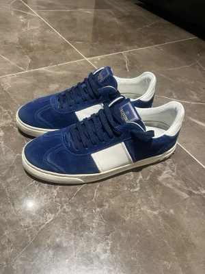 Valentino flycrews  - Snygga blå och vita sneakers från Valentino med ovandel i mocka och detaljer i vitt skinn. Klassisk låg modell med rund tå och snörning. Märkeslogga på plösen och kontrasterande vit sula. Perfekta för dig som vill ha en stilren och exklusiv look.