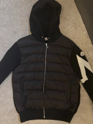 Moncler Cardigan   - Svart dunjacka från Moncler med huva och hel dragkedja. Jackan har quiltad front, stickade ärmar och en stor sopad vit blixtdetalj på ena ärmen samt Moncler-logga. Två fickor framtill med knappstängning. Är i bra skick 