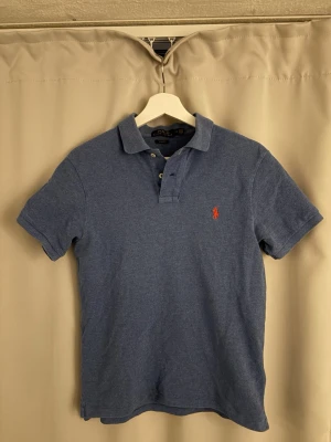 Blå pikétröja från Polo Ralph Lauren - Klassisk blå pikétröja från Polo Ralph Lauren med korta ärmar och krage. Tröjan har knappar vid halsen och den ikoniska röda logotypen broderad på bröstet. Perfekt till jeans eller shorts för en avslappnad stil. xs/s 