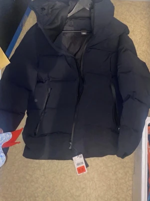 Peak performance jacka  - Snygg svart pufferjacka med huva och två snedställda dragkedjefickor framtill. Jackan har quiltad design och dragkedja hela vägen upp. Perfekt för kalla dagar och ger en modern look.