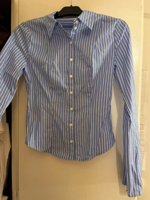 Blåvit randig skjorta med knappar - Snygg figursydd blå skjorta och klassisk krage. Skjortan har långa ärmar och knäpps framtill med vita knappar. Perfekt för en clean och stilren look. Skjortan är i storlek M. Helt ny, endast provad. Säljs pga fel storlek. 😋ps. Skulle säga att skjortan är lite mindre i storleken för att vara en M, så passar säkert S också. 