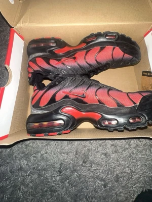 Nike Air Max Plus röd/svart sneakers - Nike Air Max Plus sneakers med ikonisk vågig design i rött och svart. Skorna har synlig Air-dämpning i sulan, mesh-överdel och svarta detaljer. Swoosh-logga på sidan och snörning för perfekt passform. Grymt snygga och sticker ut i mängden.