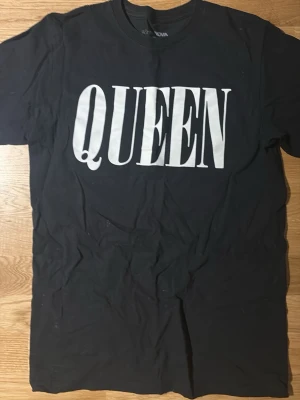 Svart Queen t-shirt med tryck - Säljer en svart t-shirt med stort vitt Queen-tryck från Fashion Nova. Klassisk passform och rund halsringning, perfekt för dig som gillar bandmerch och vill sticka ut lite extra. Använt en mycket fåtal gånger