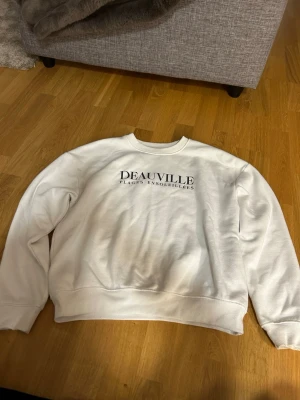 Vit sweatshirt med tryck - Vit sweatshirt med svart text 'DEAUVILLE PLAGES ENSOLEILLÉES' på bröstet. Tröjan har rund halsringning, ribbade muddar vid ärmslut och nederkant samt en avslappnad passform. 