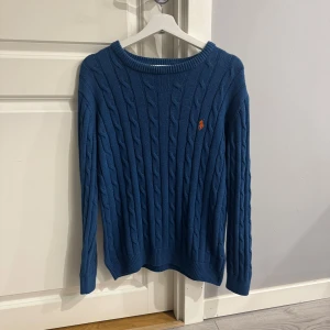 Blå kabelstickad tröja Polo Ralph Lauren - Kabelstickad blå tröja från Polo Ralph Lauren med klassisk rund halsringning och orange broderad logga på bröstet. Tröjan har lång ärm och ribbade muddar för en snygg passform. Perfekt för dig som gillar stilren och tidlös design. 