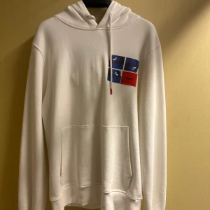 Vit hoodie från HUGO med tryck - Vit hoodie från HUGO med blått och rött tryck på bröstet samt röd patch med HUGO-logga. Tröjan har huva med dragsko, magficka och långa ärmar. Perfekt för en chill och stilren look.