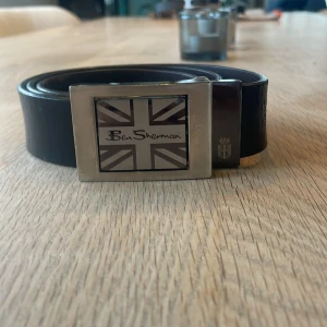 Svart skärp från Ben Sherman med metallspänne - Snyggt svart bälte från Ben Sherman med ett rektangulärt metallspänne som har Union Jack-mönster och logga. Bältet är gjort i skinn och har en stilren, klassisk form som passar till jeans eller chinos. Perfekt för dig som gillar brittisk streetstyle.