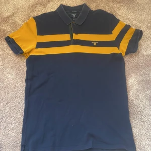 Randig GANT pikétröja i blå och gul - Snygg pikétröja från GANT i marinblå och gul med breda ränder. Klassisk krage och knappslå framtill. Tillverkad i mjuk pima bomull som känns skön mot huden. GANT-logga broderad på bröstet. Perfekt för dig som gillar sportig och stilren look. Tröjan i sig har ett bra skick med inga hål eller något, men tröjan har blivit lite uttvättat på kragen, därför säljer jag den för 200 