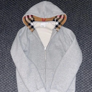 Grå Burberry hoodie med dragkedja - Säljer en grå hoodie från Burberry med klassiskt rutigt foder i huvan och broderad logga på bröstet. Tröjan har dragkedja framtill, ribbade muddar och är tillverkad i mjuk bomull. Perfekt för dig som gillar stilrena och lyxiga streetwear-plagg. Priset kan diskuteras