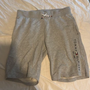 Grå shorts från Tommy Hilfiger - Snygga grå shorts från Tommy Hilfiger med logotyp och text längs benet. De har en bekväm midja med snörning i vitt och detaljer i rött och marinblått. Materialet är mjukt och stretchigt, perfekt för chill eller träning. Klassisk passform och diskret ficka bak.