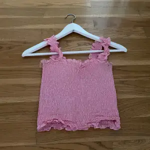 Supersöt rosa linnetopp med smockat tyg och volangdetaljer vid axelband och nederkant. 