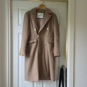 Beige kappa från Chiara Forthi, stl 36 - Snygg beige kappa från Chiara Forthi Milano med dubbelknäppning och guldiga knappar. Klassisk krage och fickor framtill. Yttertyg i 70% polyester och 30% ull, fodrad med 100% polyester. Perfekt för dig som vill ha en stilren och trendig look.