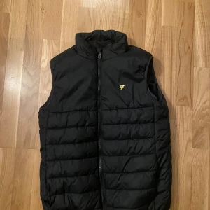 Svart väst från Lyle & Scott - Snygg svart väst från Lyle & Scott  med gul logga på bröstet. Västen har ståkrage som man kan vika ner eller ha uppe om man vill. Passar nog XS/S. Skicka gärna meddelande om ni undrar något. Nypris 800kr