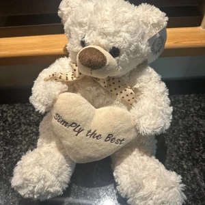 Simply the Best Teddy Bear - Mjuk och söt nallebjörn från Classic Collection, tillverkad i 100% polyester. Håller ett hjärta med texten 'Simply the Best' och har en prickig rosett. Nallen är i fint skick, med originaletikett kvar. Perfekt som present eller dekoration.