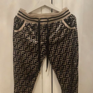 Fendi mönstrade mjukisbyxor med snörning - Snygga mjukisbyxor från Fendi med ikoniskt FF-mönster i svart och guld. Byxorna har bred resår med svarta ränder, snörning i midjan och fickor. Materialet är mjukt och glansigt, perfekt för en trendig streetstyle-look.