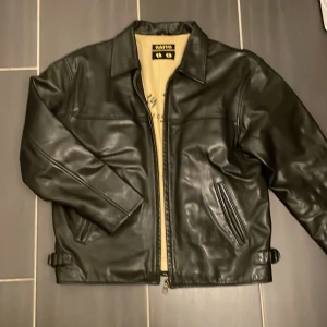 Svart skinnjacka från Gatto - Snygg svart skinnjacka från Gatto med klassisk krage, dragkedja framtill och justerbara spännen i sidorna. Insidan är beige med broderad text 'Rusty Dusty 1956'. Jackan har raka ärmar och två fickor med dragkedja. Perfekt för dig som vill ha en tidlös och cool look.