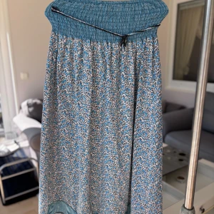 Blommig plagg med blå detaljer (används som både kjol och sommarklänning) - Säljer en långklänning/kjol med blommigt mönster i blått, grått och vitt. Överdelen är smockad med blå och svarta ränder och har en dekorativ snörning med ringdetalj. Kjoldelen är luftig och avslutas med en randig blå kant.