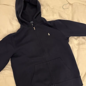Mörkblå hoodie från Polo Ralph Lauren - Säljer en mörkblå hoodie från Polo Ralph Lauren i storlek S. Tröjan har dragkedja framtill, huva och en klassisk känguruficka. Ikoniska Polo-loggan broderad på bröstet. Perfekt för chill dagar och snygg streetstil. Barnstorlek XL eller vanlig S. Som ny kvalitet!