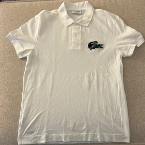 Vit Lacoste pikétröja i bomull - Vit pikétröja från Lacoste med klassisk krage och två knappar framtill. Ikonisk krokodillogga broderad på bröstet. Tillverkad i bomull med regular fit. Perfekt för dig som gillar stilren och sportig look.