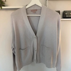 Beige kofta med fickor och knappar - Snygg beige kofta med v-ringad hals och stora knappar framtill. Koftan har två praktiska fickor och ribbade detaljer vid ärmslut och nederkant. Perfekt för lager-på-lager och chill dagar. Passar dig som gillar enkel och clean stil.