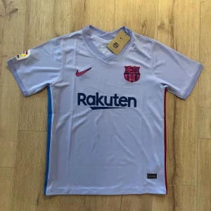 Barcelona fotbollströja Nike M - Snygg Barcelona fotbollströja från Nike i ljuslila med mörkblå och röda detaljer. Tröjan har korta ärmar, V-ringad krage och är tillverkad i Dri-FIT material som andas. FCB-emblem på bröstet, La Liga-märke på ärmen och Rakuten samt UNICEF tryck. Perfekt för dig som älskar Barca!