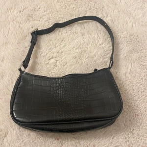 Svart axelväska från Monki - Snygg svart axelväska från Monki med croco-mönstrad yta och justerbar rem. Väskan har en stilren, avlång form och stängs med dragkedja upptill. Perfekt för dig som gillar trendiga accessoarer med lite edge.