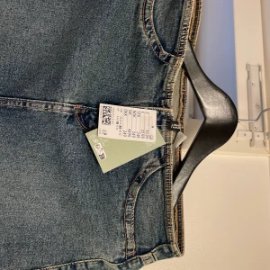 Ny lång jeanskjol från H&M med slits - Säljer en snygg lång jeanskjol från H&M i klassisk blå denim. Kjolen har en rak passform, framfickor och bakfickor samt en cool slits framtill som ger extra edge. Perfekt att styla med sneakers eller boots för en chill vibe. Kjolen är ny med prislappen.