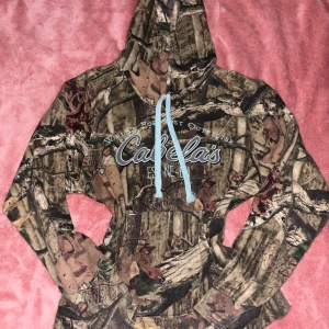 Camo hoodie - Säljer en hoodie från Cabela's i storlek L med camouflagemönster och ljusblå snörning. Tröjan har stor huva, magficka och tryckt logga på bröstet. Insidan är mjuk på insidan för extra komfort. Perfekt för dig som gillar t.ex streetstyle och baggy passform! Skriv gärna innan köp💕