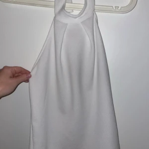 Vit halterneck topp med ringdetalj - Supersnygg vit halternecktopp med cool ringdetalj vid halsen. Toppen har en slät framsida och insida i satinliknande material som känns lyxigt mot huden. Perfekt för dig som gillar minimalistisk stil och vill sticka ut med en unik detalj.