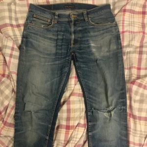 Blå jeans från Nudie Jeans Co - Säljer ett par klassiska blå jeans från Nudie Jeans Co med slitningar och hål på knäna. Jeansen har fem fickor, orangea sömmar och en snygg tvättad look. Perfekt för dig som gillar en avslappnad och trendig stil.