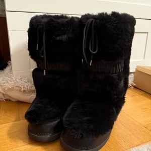 Svarta pälsboots från Moschino - Säljer ett par svarta fluffiga boots från Moschino med pälsimitation och snörning framtill. Snygg logga runt skaftet och röda hjärtan med Love Moschino-text på hälen. Perfekta för vintern och riktigt statement med sin chunky look.
