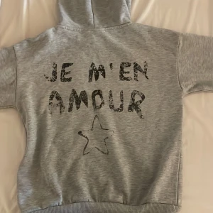 Grå hoodie med tryck från Gina Tricot - Säljer en grå hoodie från Gina Tricot med svart texttryck 'JE M'EN AMOUR' och en stjärna på både fram- och baksidan. Tröjan har huva, långa ärmar och ribbade muddar. 