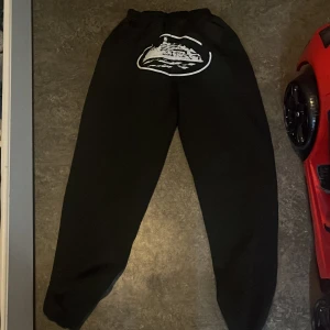 Svarta cortez mjukisbyxor - Svarta mjukisbyxor med elastisk midja och ett stort vitt tryck av ett skepp på baksidan. Byxorna har en loose passform och är tillverkade i mjukt material som är perfekt för chill dagar. Snyggt och enkelt designade för en avslappnad stil.