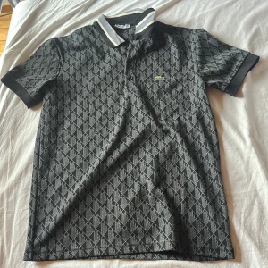 Svart mönstrad Lacoste piké S - Svart pikétröja från Lacoste i storlek S med grått geometriskt mönster över hela plagget. Klassisk krage med vita och grå ränder, knappar med Lacoste-logga och den ikoniska gröna krokodilen broderad på bröstet. Perfekt för dig som gillar sportig och stilren vibe.