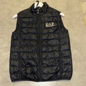 Svart dunväst från EA7 Emporio Armani - Svart dunväst från EA7 Emporio Armani med quiltad design och hög krage. Västen har dragkedja framtill och logga broderad på bröstet. Lätt och smidig modell som passar perfekt till lager-på-lager-stil.