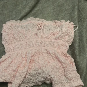 Ljusrosa spets tubtopp från Gina Tricot - Supersöt ljusrosa tubtopp från Gina Tricot med broderad spets och smockad resår. Toppen har en liten rosett framtill och volangkant upptill och nertill. Perfekt för varma dagar och ger en romantisk vibe.