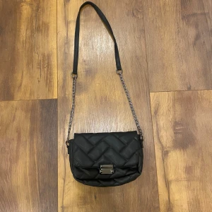 Svart quiltad axelväska med kedja - Snygg svart axelväska med quiltad design och silverfärgad kedja. Väskan har ett stilrent spänne framtill och axelrem i både kedja och skinnimitation. Perfekt för dig som vill ha en trendig och praktisk väska med det lilla extra.