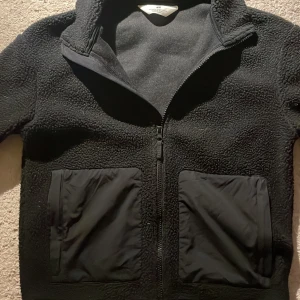 Svart teddyjacka från H&M med dragkedja - Svart teddyjacka från H&M med hel dragkedja framtill och två stora fickor, säljer pga av inte kommer till användning!