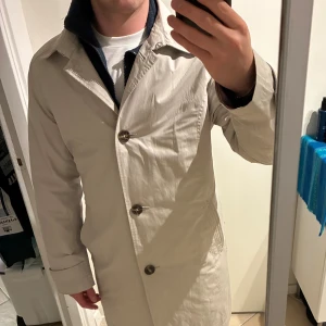 Beige trenchcoat från Saki, stl 42 - Stilren beige trenchcoat från Saki med klassisk krage, knäppning med stora knappar och bälte i midjan. Jackan har långa ärmar och är tillverkad i ett slätt material som känns lätt och fräscht. Perfekt för dig som gillar en clean och tidlös look. Storlek 42/XS